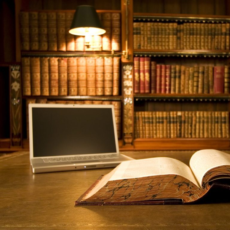 Ein offenes Buch liegt auf einem Tisch neben einem Laptop und einer Bücherwand.